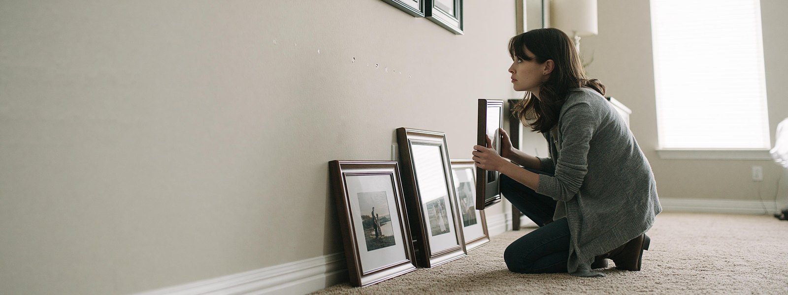 Woman crouched on a carpet beside three framed pictures leaning against her living room baseboard, one frame in her hands, looking up at a short row of small primed spots on the finished wall where the hooks came out