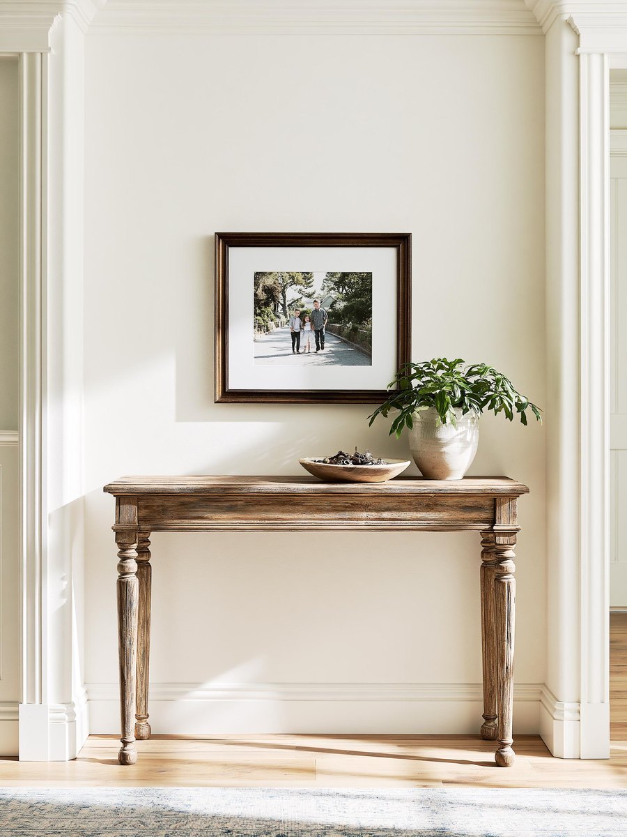 Single framed photograph hung back on a finished entry wall painted a warm off white, the wall reading as one even surface behind it, a narrow console table below with a bowl of keys, bright even daylight crossing the wall