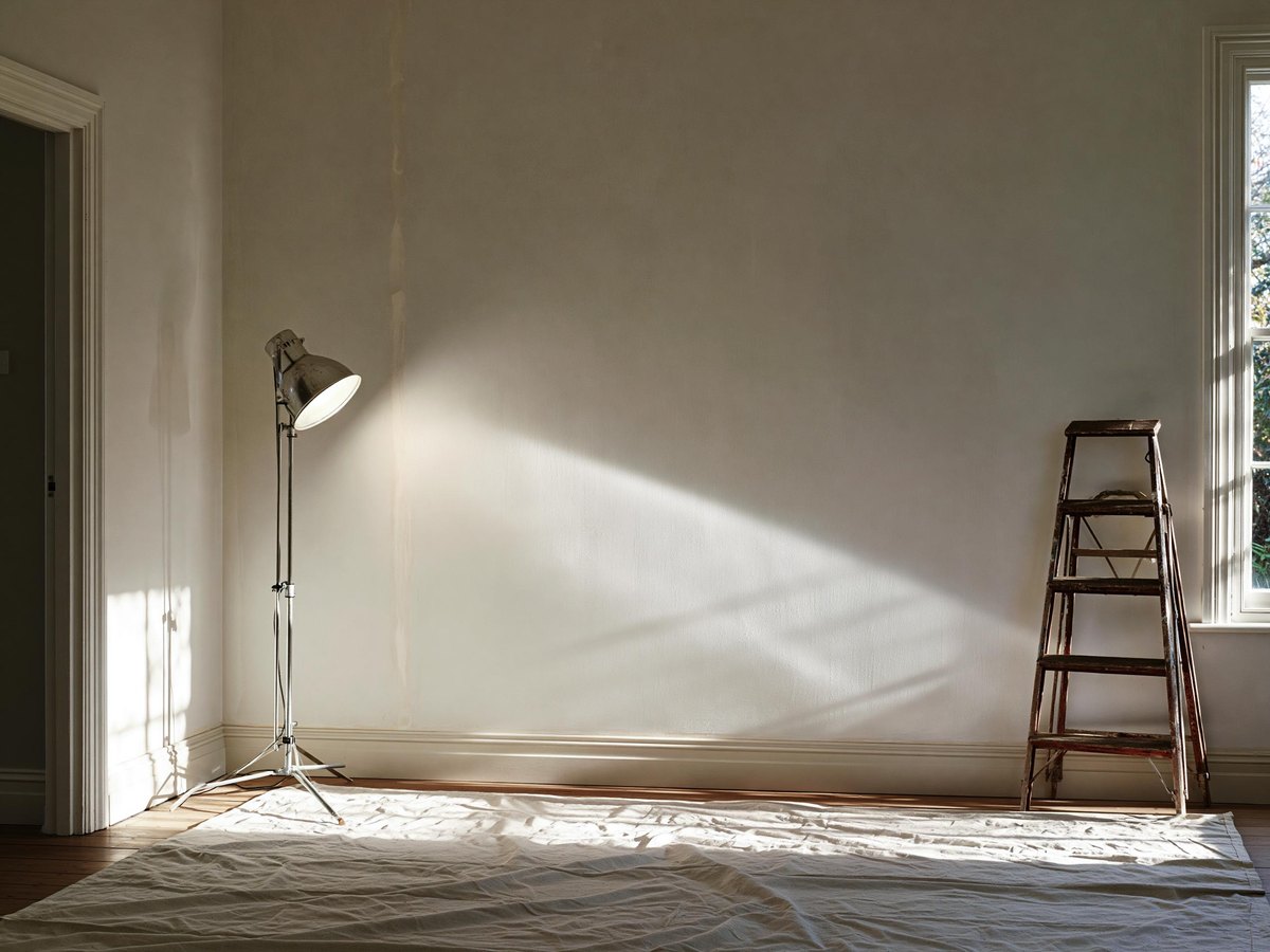 Work light on a low stand set close in to a finished wall so its beam skims across the surface, one taped joint picked out as a faint pale band running floor to ceiling, a folded step ladder against the far wall