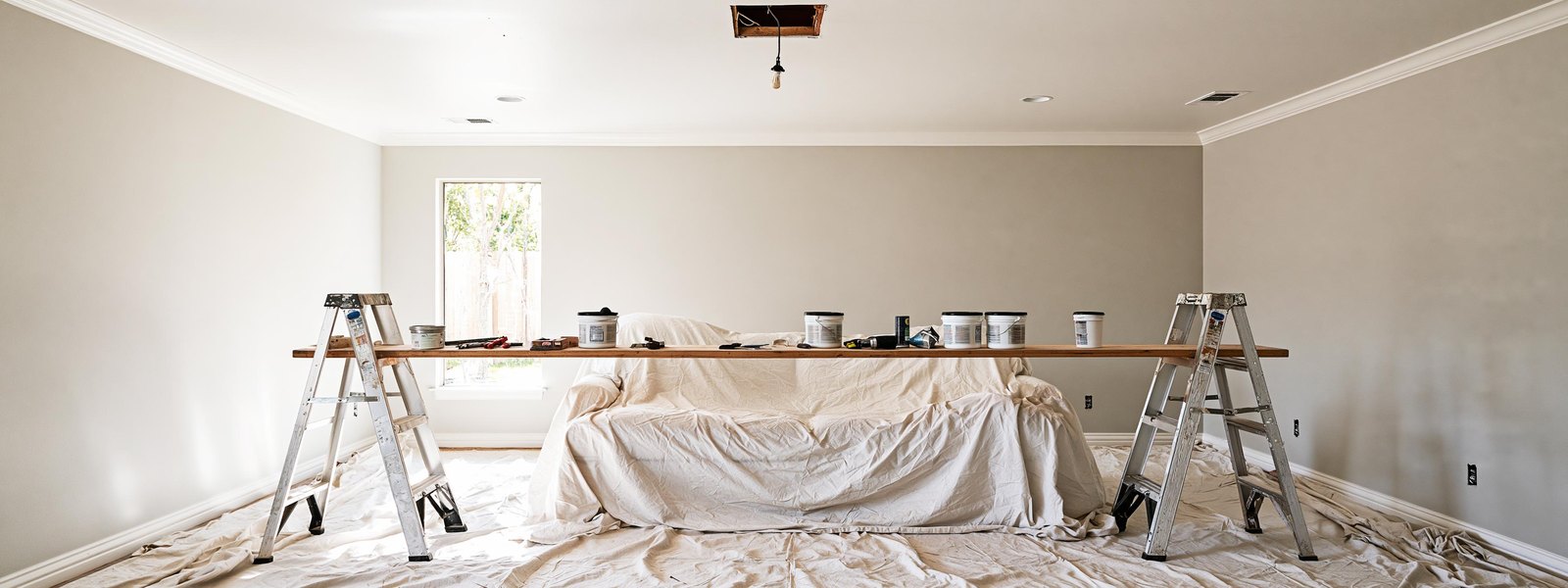 Working plank set level between two step ladders across a furnished living room, canvas drop cloths over the floor and the sheeted sofa below, the ceiling above finished flat white and even with the light fixture taken down and its box left open